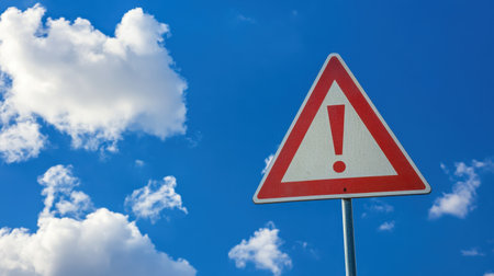 A red triangular warning sign with an exclamation mark stands tall against a vibrant blue sky dotted with white clouds, serving as a symbol of caution in various environments.の素材