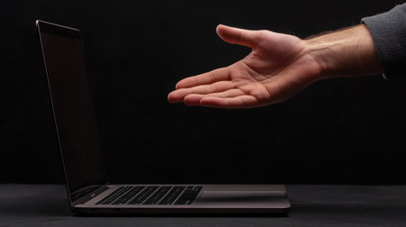 A human hand reaches towards an open laptop on a dark background, representing modern technology and virtual interaction in a minimalist workspace.の素材