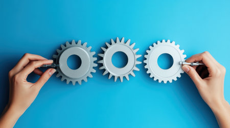 Close-up view of hands assembling gray gears against a vibrant blue background, symbolizing innovation and teamwork in engineering and design processes.の素材