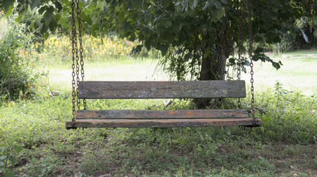A rustic wooden swing hangs from a tree in a lush green landscape. This tranquil setting evokes childhood memories and a sense of peaceful relaxation in nature.の素材