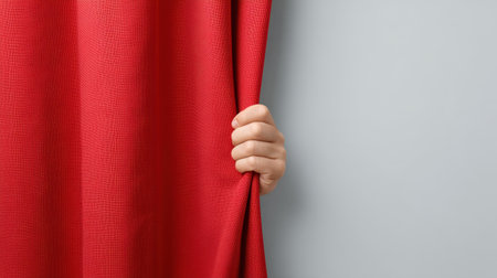 A close-up view of a hand holding a red curtain, set against a gray wall, symbolizing anticipation and excitement before a theater performance.の素材