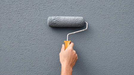 A person using a paint roller to apply gray paint on an exterior wall, showcasing the effort in home improvement and emphasizing a fresh, modern look.の素材
