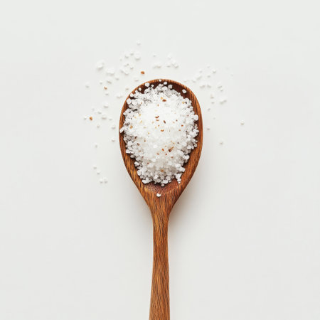 A close-up image of coarse sea salt in a wooden spoon, with granules scattered on a clean white background, ideal for culinary, cooking, and food styling purposes.の素材