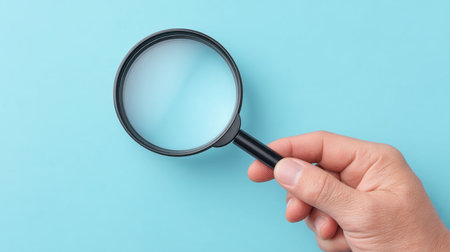 A hand holds a magnifying glass against a light blue background, symbolizing investigation and observation. Ideal for educational and research themes.の素材