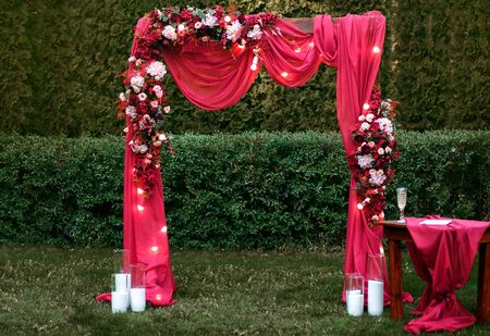 Wedding ceremony, wedding arch, red wedding arch of flowers and greenery stands on the green grass in the park, beautiful color, original composition, table with glasses of champagneの写真素材