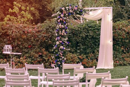 Wedding. Ceremony. Wedding arch. A wedding arch of flowers and greenery stands on the green grass in the park, a beautiful sunset, white chairs, the beginning of the wedding ceremonyの写真素材