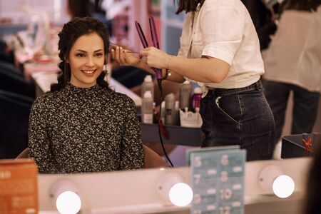 young woman in the women's salon uses the services of a hairdresser, a professional hairdresser straightens her hair with a curling iron, the girl smiles and is satisfied with the work of the stylistの写真素材