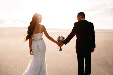 The bride and groom in the desert, holding a wedding bouquet in their hands, love and tenderness in the dunes of the desert, a beautiful sunset, a beautiful woman looks at her beloved manの写真素材