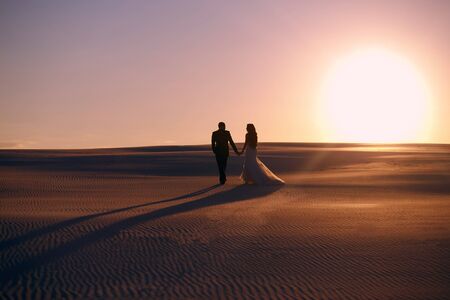 bride and groom walk in the desert during sunset, the silhouette of a man and woman, a man leads a girl along the dunes of the desert, a wedding walk, the concept of a family overcoming difficultiesの写真素材