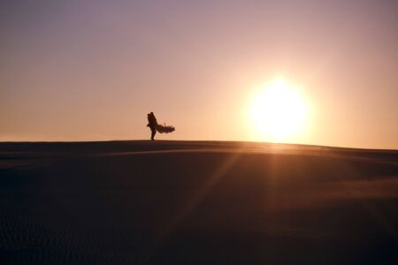 The bride and groom walk in the desert during sunset, a man holds a woman in his arms, people silhouettesの写真素材