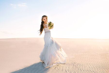 beautiful bride in a white wedding dress walks in the sandy desertの写真素材
