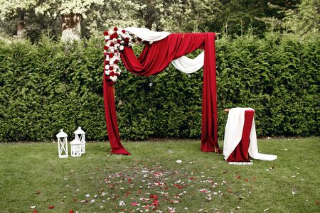 The wedding arch is decorated with red-white fabrics and fresh flowers, the wedding ceremony in the garden with a sheared lawnの写真素材