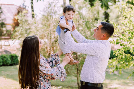 Father and mother are holding their little son in arms, having fun on a green lawn