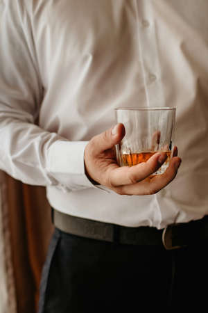 in the office there is a man with a glass of whiskey in a white shirt, vertical picture, male mood, focus on alcohol, relax after a hard day, strong arm, bourbon in a stressful situationの写真素材