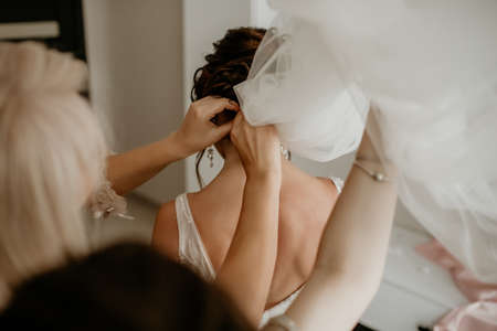 wedding day, girlfriends help the bride to wear a dress and veil, a young bride is waiting for the...