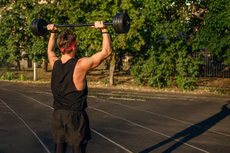Man athlete lifts the barbell over his head, training athlete outdoors at sunsetの写真素材