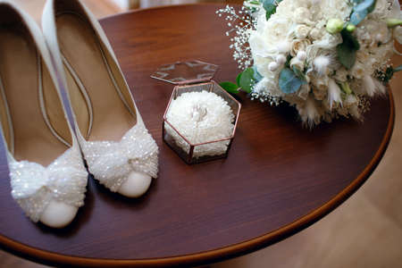 Wedding accessories in the form of a bouquet, rings and shoes are on a wooden table.