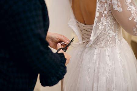 the bride tries on a wedding dress before buying, the seamstress cuts the long ribbons with scissorsの写真素材