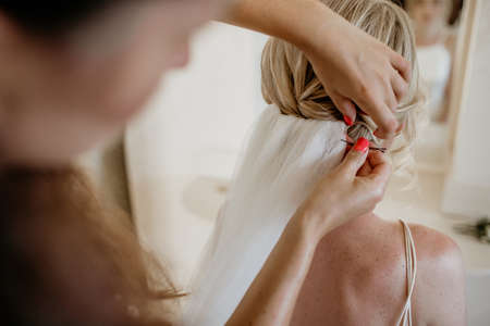 A professional hairdresser does a wedding hairstyle and throws a white veil over a young beautiful blondeの写真素材