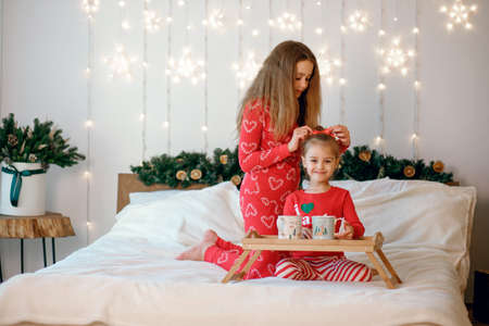New Year and Christmas holidays in a beautifully decorated room. Two sisters are sitting on the bed in red pajamas and braiding each others hairの写真素材