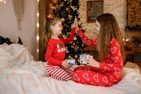 Two cute little sisters in red pajamas sit on the bed and cheerfully exchange giftsの写真素材