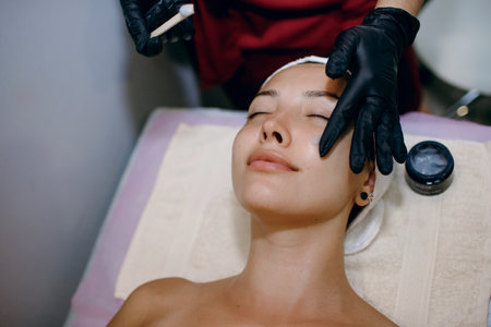 A beautiful brunette is undergoing treatments at the spa, a master beautician applies a moisturizer...
