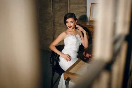 Beautiful woman bride with perfect hair and manicure sits in a wedding dress and poses looking at the photographerの写真素材