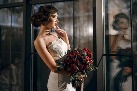 A beautiful girl bride in a white dress and white gloves with a huge bouquet of roses stands against the background of a wooden wall and windowの写真素材