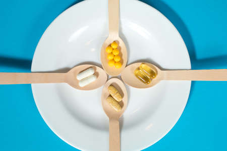 Vitamins on wooden spoons in a white plate on a blue background. Multivitamin set for every day. The importance of obtaining a vitamin complex to improve the functioning of the body and prevent healthの写真素材