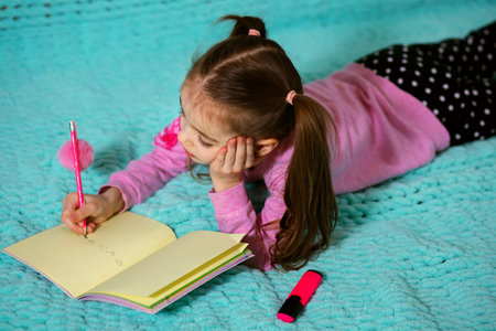A little girl in a pink sweater with pigtails lies on a blue sofa and draws in a notebook with a pink pencilの写真素材