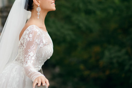 Happy beautiful bride in a white wedding dress smiles and holds a handrail on a background of green leavesの写真素材
