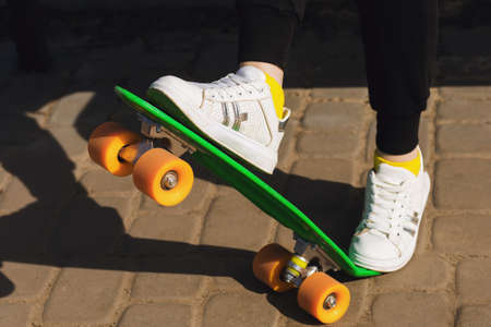 A child in white sneakers rides a green skateboard or pennyboard. The girl learns to perform tricks on a skateboard. Skateboarding is a fashionable means of transportation and a great fitness toolの写真素材