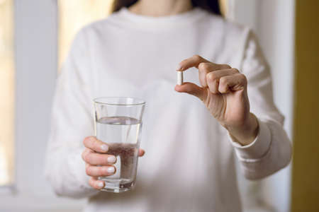 A woman holds a glass of water and a pill in her hands close-up. Health problems and treatment with...