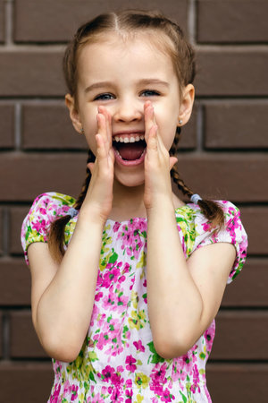 Baby girl screams loudly, holding her hands to her mouth. A preschool girl stands by a brick wall...