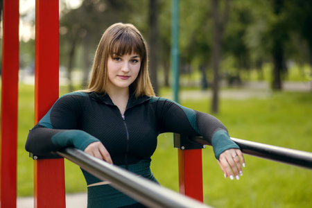 An athletic young woman in sportswear stands on an open sports ground with horizontal bars designed for training. Strong brunette goes in for sports increasing the volume of musclesの写真素材