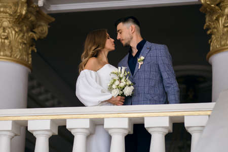 The bride and groom stand indoors and hug looking at each other. A beautiful bride holds a bouquet of flowers and is dressed in a luxurious wedding dress. Stylish groom getting ready to kiss his brideの写真素材