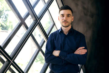 A young man in a blue shirt stands indoors by the window and looks confidently. A stylish guy with a beard crossed his arms over his chest. Model man posing for photographerの写真素材