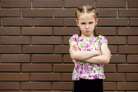The little girl is very unhappy and pouted into a closed pose. A child with braided hair and a bright T-shirt against a brick wall is upset and not in a good moodの写真素材
