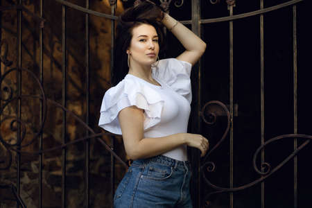 Portrait brunette woman on the background of a wrought iron fence at sunset. The girl is dressed in a white T-shirt and denim shorts. Beautiful woman straightens her hair and poses for a photographerの写真素材