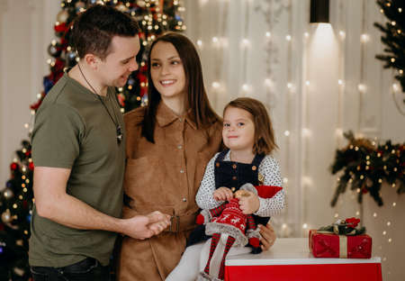 Young family dad, mom and daughter celebrate christmas and new year at home. Happy family rejoice and enjoy the family holiday on the background of a Christmas tree with garlands and giftsの写真素材