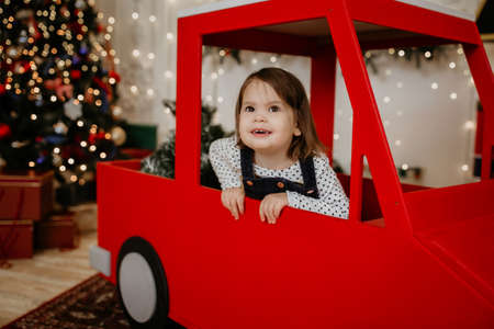 Little girl child sits in a red toy car on the background of a Christmas tree and smiles. New Year and Christmas holidays. Happy childhood and family celebration. Christmas tree with garlandsの写真素材