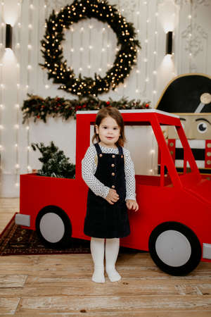 A little girl stands near a red toy car against the background of a Christmas wreath made of spruce. New Year and Christmas holidays. Happy childhood and family holiday. Christmas tree with garlandsの写真素材