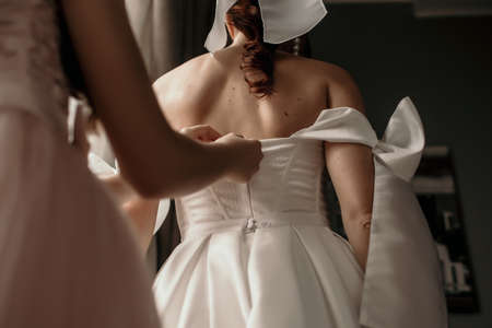 A bride wears a white wedding dress in a hotel room. The bridesmaid or mom helps to lace up the bride's wedding dress. Happy newlyweds on their wedding dayの写真素材