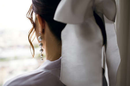 A girl in a white robe looks out the window. A woman with a white hair bow and pearl earrings looks out the window of her hotel. Wedding morning of the brideの写真素材
