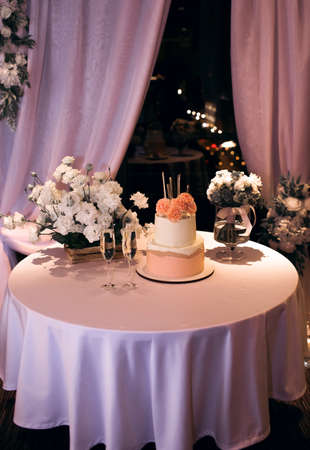 Wedding cake with glasses of champagne on the table, decorated with fresh white flowers. Wedding evening with dessert in the form of a beautiful cake with candles. Traditional wedding ceremonyの写真素材