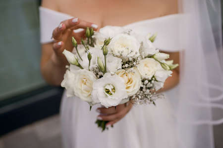 The bride in a wedding dress and a long veil holds a white wedding bouquet of roses close-up. A beautiful woman bride examines and touches her wedding bouquet. The girl is wearing wedding ringsの写真素材