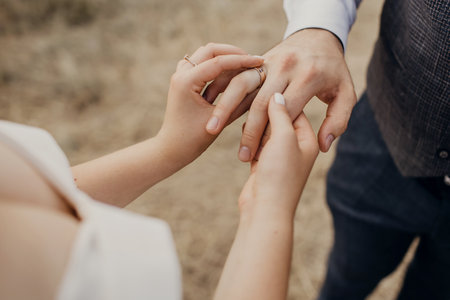 The bride puts the wedding engagement ring on the groom's finger at the wedding ceremony. Hands of newlyweds with rings close-up. Heterosexual young couple in love dressed in stylish wedding clothesの写真素材