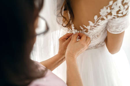 Mom helps her daughter, the bride, button up her white wedding dress on her wedding day. Hands and buttons close up. The affectionate relationship between mother and daughterの写真素材