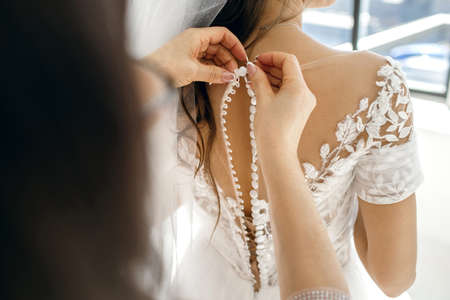 The bridesmaid helps the bride button up her white wedding dress on her wedding day. Hands and buttons close up. An affectionate relationship between mother and daughterの写真素材