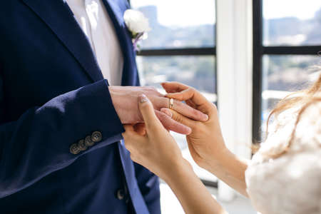 In an indoor wedding ceremony, the bride puts the wedding ring on the groom's finger, side view....